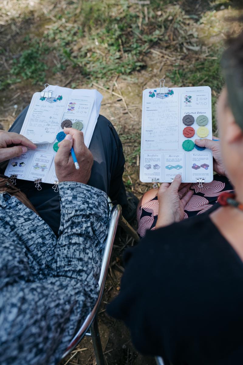 Zwei Personen spielen im Freien ein Rollenspiel mit Notizbögen und bunten Spielsteinen