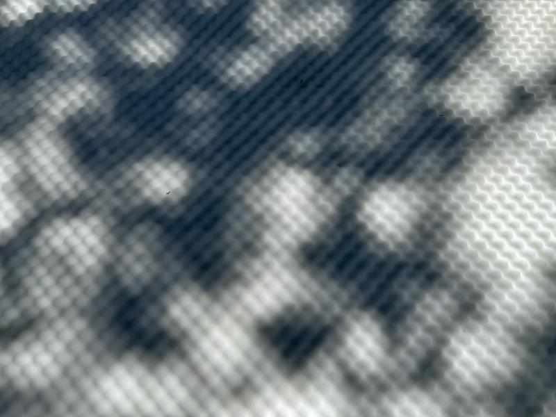 Licht und Schatten werfen ein rasterartiges Muster auf eine strukturierte Oberfläche und erzeugen ein abstraktes Spiel aus Formen und Tiefen.