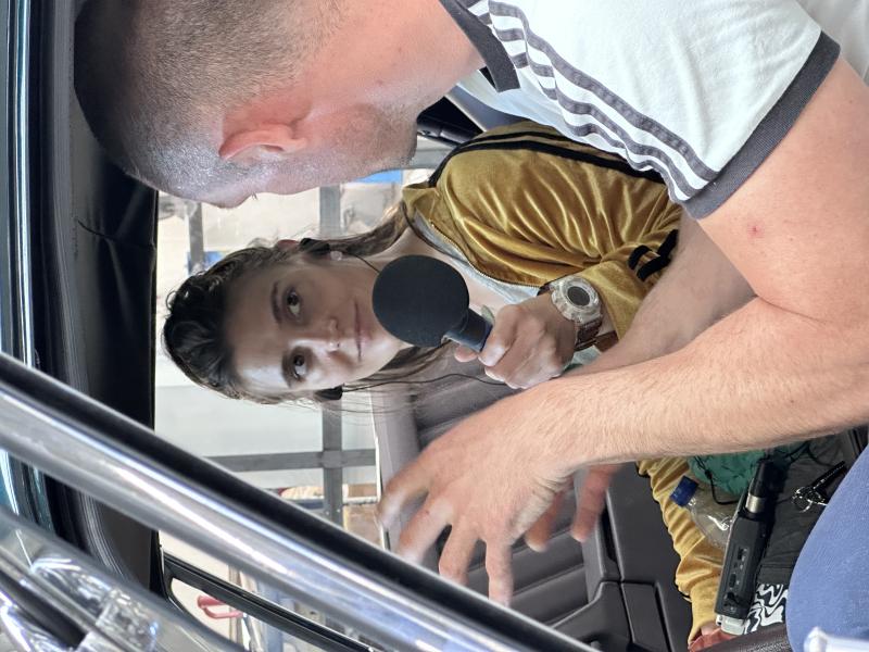 Eine Frau hält ein Mikrofon und interviewt einen Mann in einem Auto, während beide ein Gespräch führen.