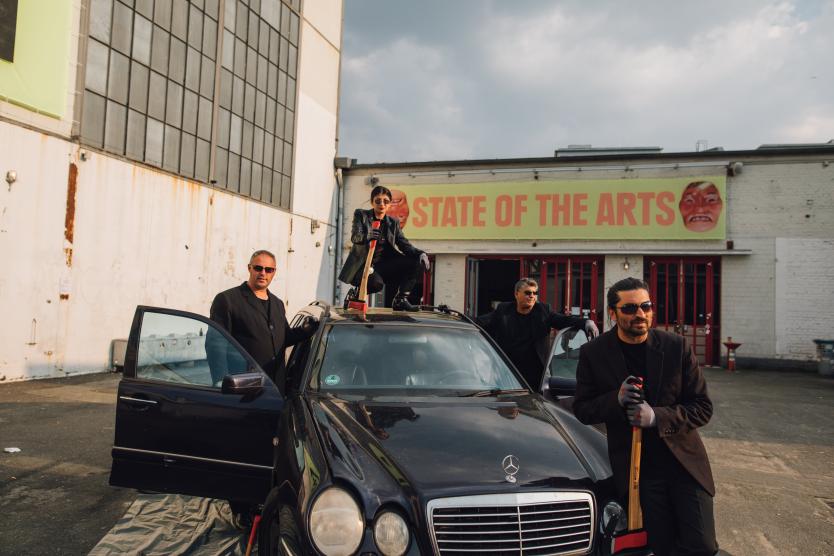 Das Bild zeigt eine Gruppe von vier Personen in schwarzer Kleidung, die um ein schwarzes Auto herum posieren, während eine Person mit einer Axt auf dem Dach des Fahrzeugs steht, vor einem Gebäude mit der Aufschrift „STATE OF THE ARTS“.