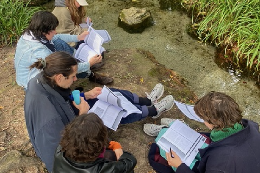 The image shows a group of five people sitting by the bank of a small stream in the forest, immersed in books and notes, while surrounded by dense vegetation.