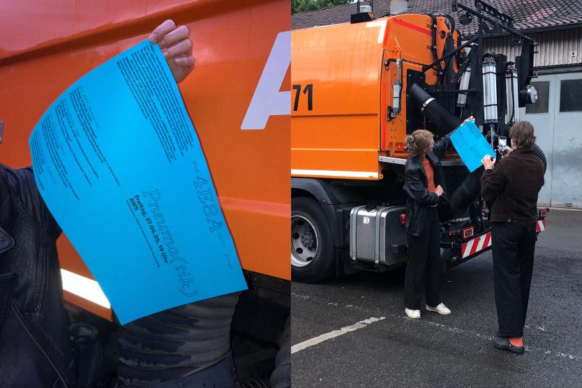 Zwei Personen präsentieren ein blaues Plakat vor einem orangefarbenen städtischen Reinigungsfahrzeug, während eine von ihnen es fotografiert.