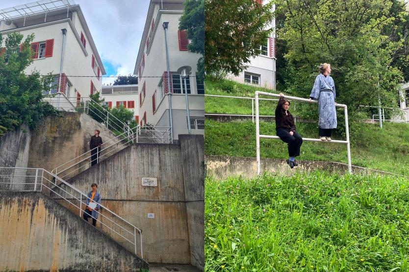 Links zwei Personen auf einer steilen Außentreppe zwischen weißen Gebäuden mit roten Fensterläden, rechts zwei weitere Personen an einem Geländer auf einer grünen Böschung vor ähnlicher Architektur.