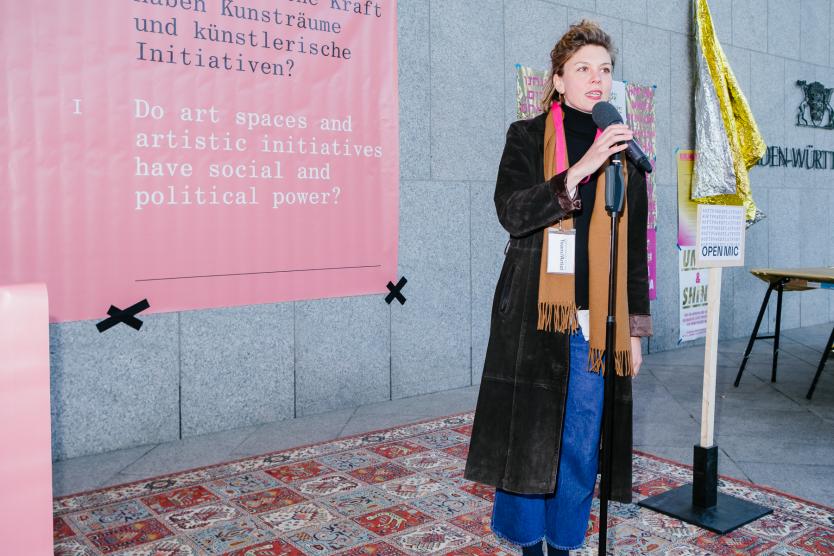 Eine Frau spricht bei einer öffentlichen Veranstaltung ins Mikrofon, hinter ihr ein Banner mit politischer Botschaft.