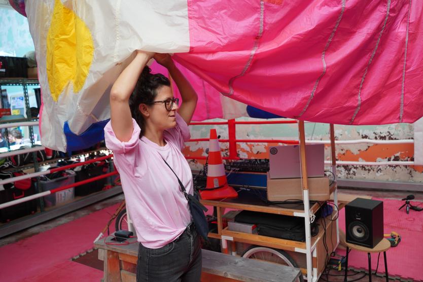 Eine Frau in einem pinken Raum richtet ein großes, buntes Stofftuch über einem mobilen DJ-Pult aus.
