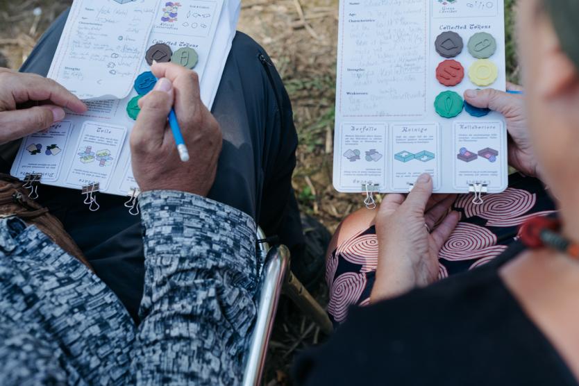 Zwei Personen spielen im Freien ein Rollenspiel mit Notizbögen und bunten Spielsteinen