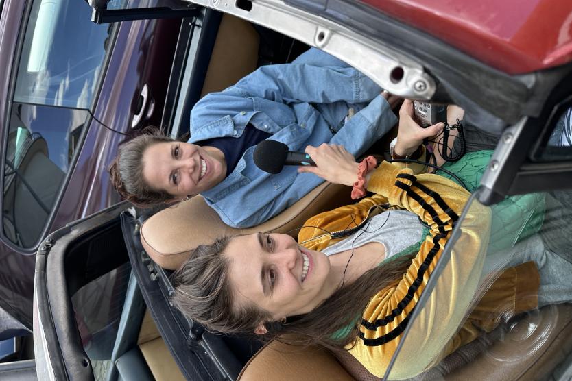 Zwei Frauen sitzen in einem Cabrio und führen ein Interview, bei dem eine der beiden ein Mikrofon hält und der anderen aufmerksam zuhört.