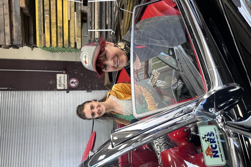 Zwei Frauen sitzen in einem klassischen Cabrio mit rotem Innenraum, lächeln in die Kamera – eine Mischung aus Interview und entspannter Oldtimer-Atmosphäre in einer Werkhalle.