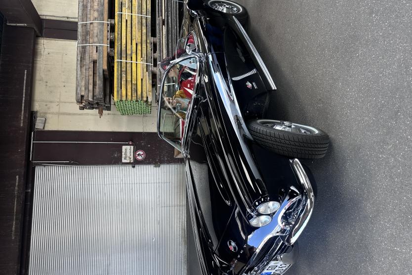 Zwei Frauen sitzen in einem schwarzen Oldtimer-Cabrio, während sie ein Interview führen – die Szene spielt sich in einer Lagerhalle mit industriellem Charme ab.