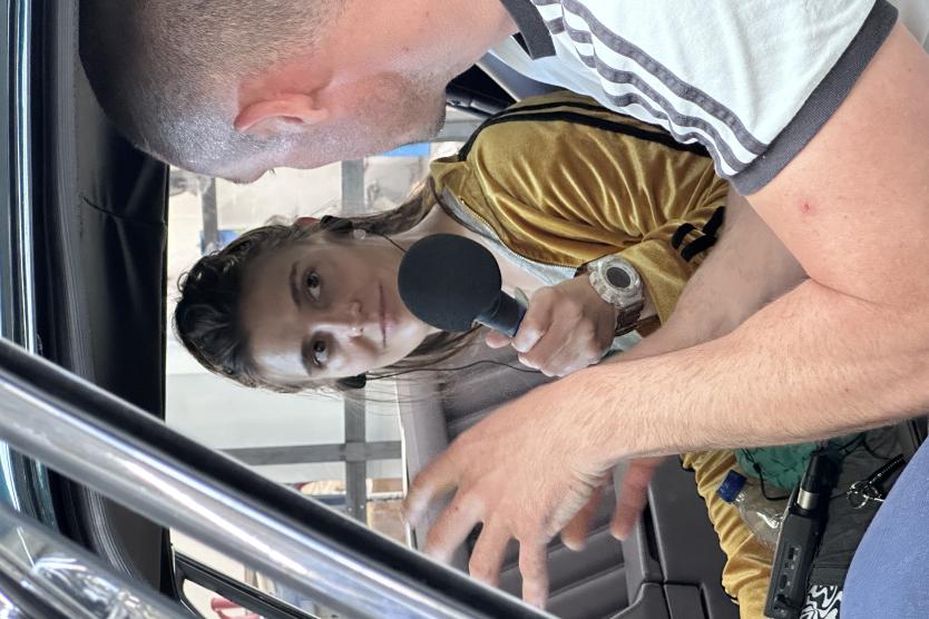 Eine Frau hält ein Mikrofon und interviewt einen Mann in einem Auto, während beide ein Gespräch führen.