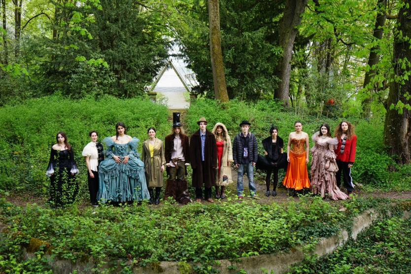 Eine Gruppe von elf Personen steht aufgereiht in einer parkähnlichen Naturkulisse und trägt aufwendige, teils historische Kostüme und Hüte.   A group of eleven people stands lined up in a park-like setting, wearing elaborate, partly historical costumes and hats. 
