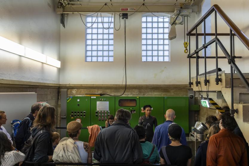 Eine Gruppe von Zuschauern steht in einem Industrie- oder Pumpenraum vor einer Performerin, im Hintergrund sind grüne Schaltschränke und Glasbausteinfenster zu sehen.   A group of spectators stands in an industrial or pump room facing a female performer. Green control cabinets and glass-block windows are visible in the background. 