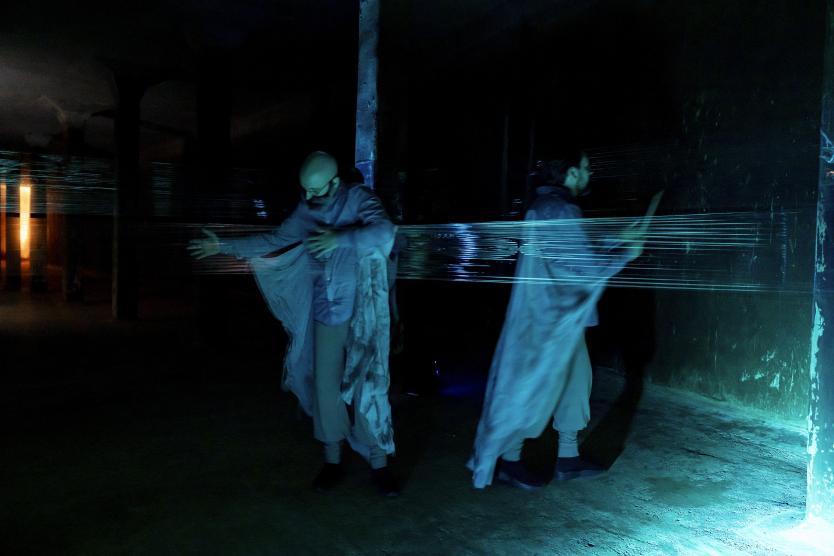 Zwei Performer in bläulichem Licht bewegen sich hinter einer gespannten, transparenten Folienbahn in einem dunklen Raum. Two performers move in bluish light behind a stretched, transparent sheet of plastic in a dark room. 