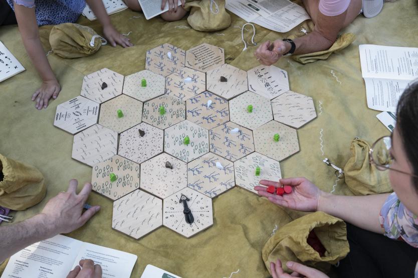 Draufsicht auf ein Brettspiel aus sechseckigen Feldern und Spielfiguren, das von mehreren Personen im Freien gespielt wird.   Top-down view of a board game made of hexagonal tiles and game pieces being played outdoors by multiple people. 