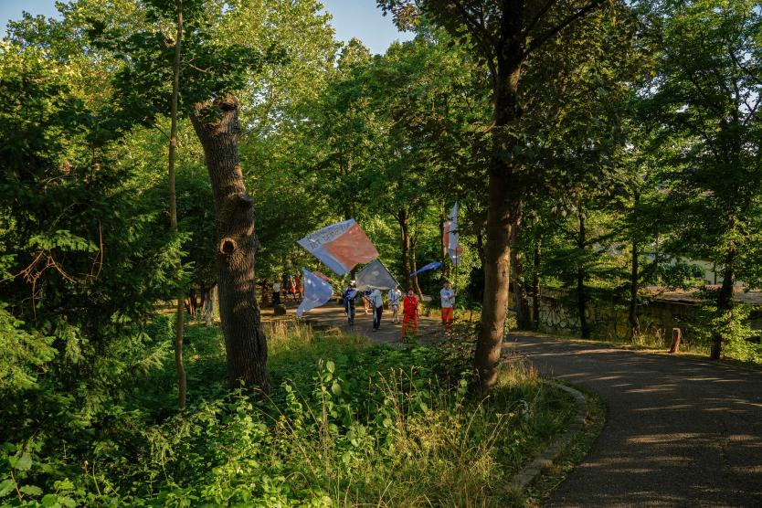 Eine Gruppe von Menschen zieht mit farbigen Fahnen über einen bewaldeten Parkweg bei sonnigem Licht.   A group of people walks along a wooded park path in sunny light, carrying colorful flags. 