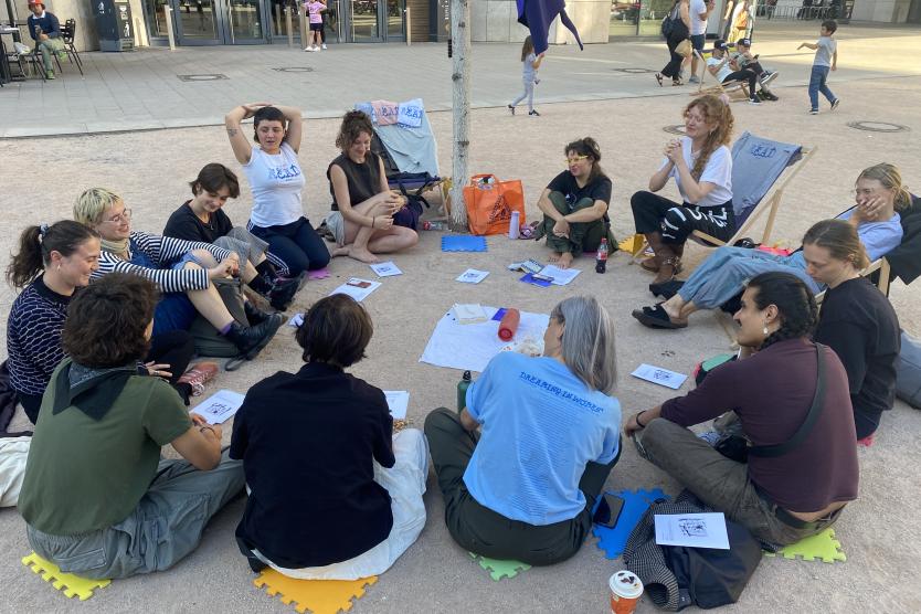 Eine Gruppe von Menschen sitzt im Kreis auf bunten Unterlagen auf einem gepflasterten Platz im Freien und tauscht sich aus.   A group of people sits in a circle on colorful pads in a paved outdoor plaza, engaged in conversation. 