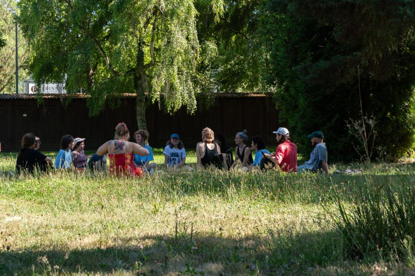 Eine Gruppe von Menschen sitzt im Kreis auf einer schattigen Wiese unter Bäumen zusammen.  A group of people sits together in a circle on a shady lawn beneath tall trees. 