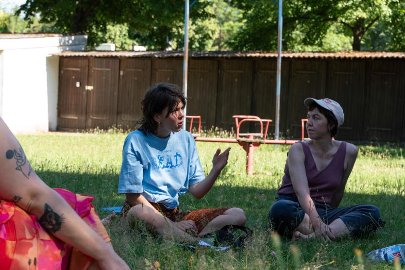 Zwei Frauen sitzen im Gras während einer Gesprächsrunde im Freien; eine im blauen T-Shirt gestikuliert beim Sprechen.  Two women sit in the grass during an outdoor discussion; one wearing a blue t-shirt gestures while speaking. 