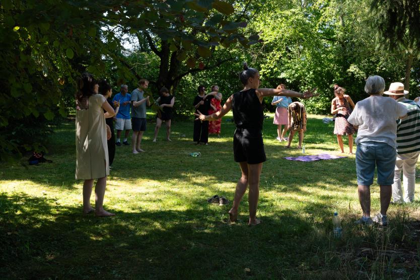 Eine Gruppe von Menschen steht auf einer sonnenbeschienenen Lichtung im Park und führt gemeinsam Bewegungs- und Atemübungen durch.   A group of people stands in a sunlit park clearing, practicing movement and breathing exercises together. 
