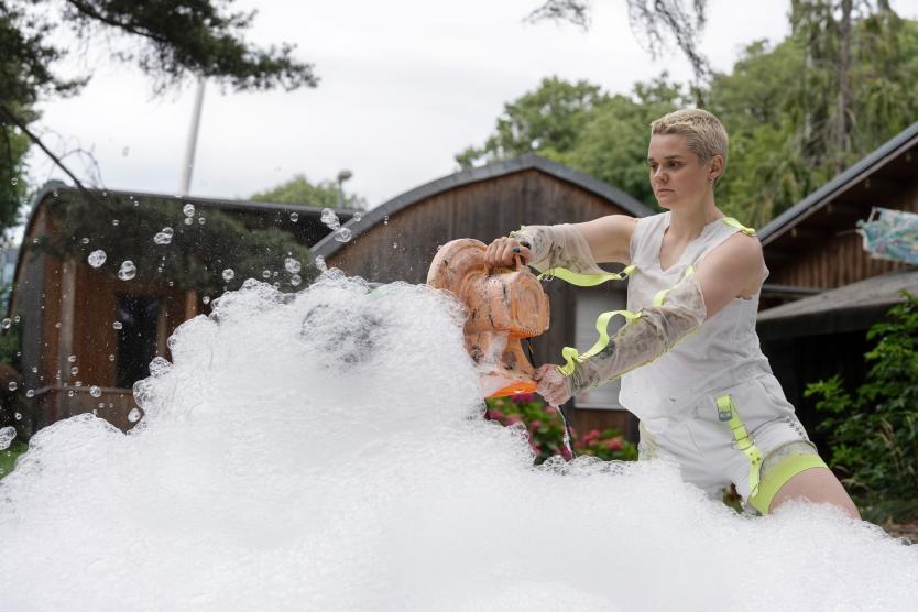 Eine Künstlerin in weißer Kleidung bedient ein oranges Gebläse, das große Mengen weißen Schaum erzeugt.  An artist in white clothing operates an orange blower machine that produces large quantities of white foam.
